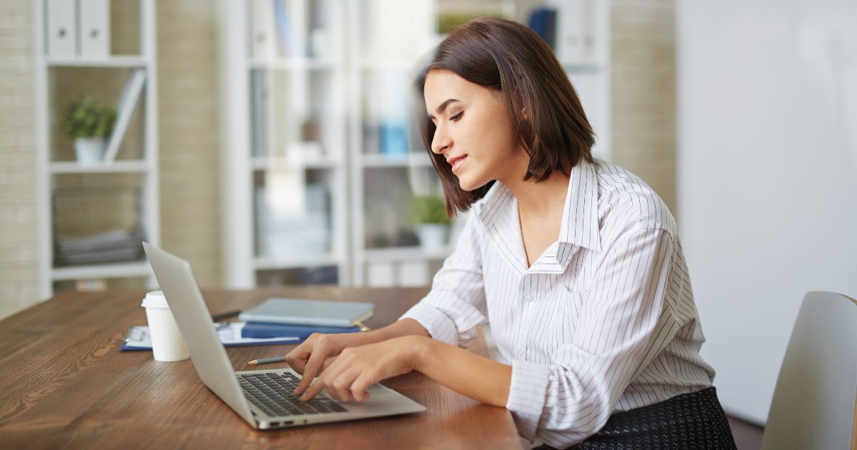 Young woman working on a laptop in a modern office setting, focused on her tasks, with a coffee cup and notebooks on the wooden table.
